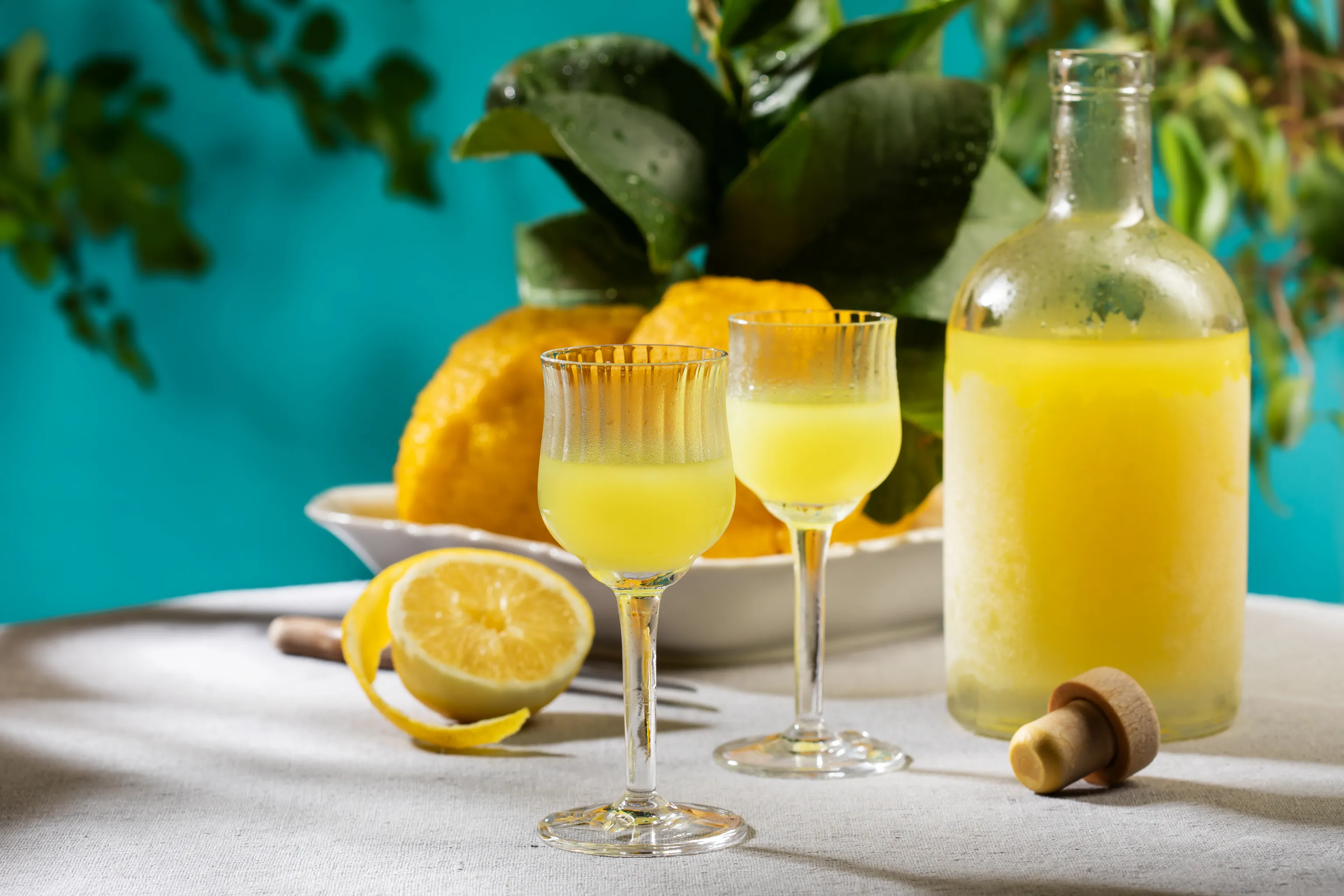 Zwei Gläser mit gelbem Likör, eine Flasche und frische Zitronen auf einem Tisch vor einem leuchtend blauen Hintergrund und grünem Laub.