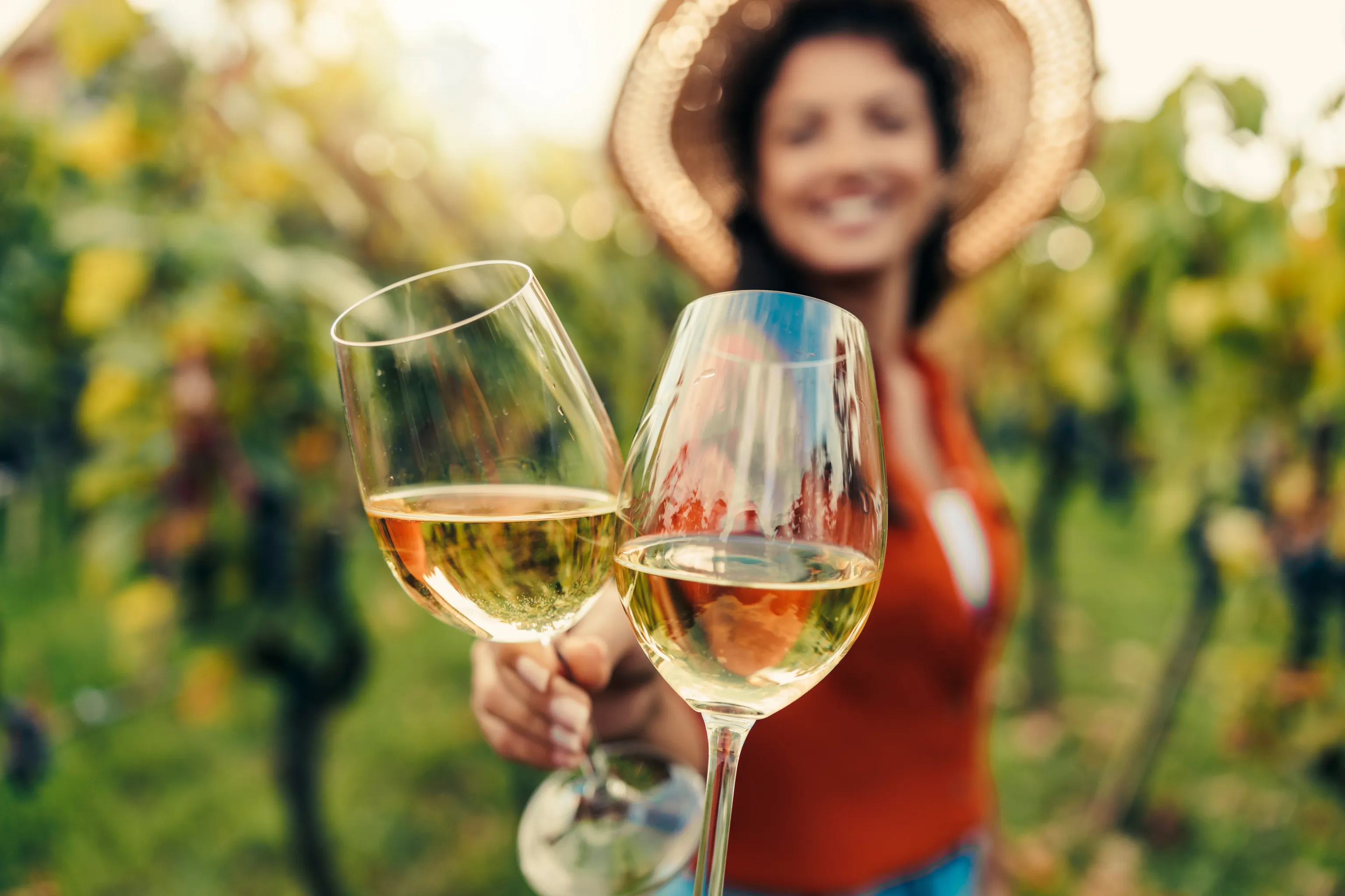 Zwei Gläser Weißwein, die zum Anstoßen erhoben werden, im Hintergrund eine lächelnde Person mit Sonnenhut, die im Grün der Weinberge verschwimmt.