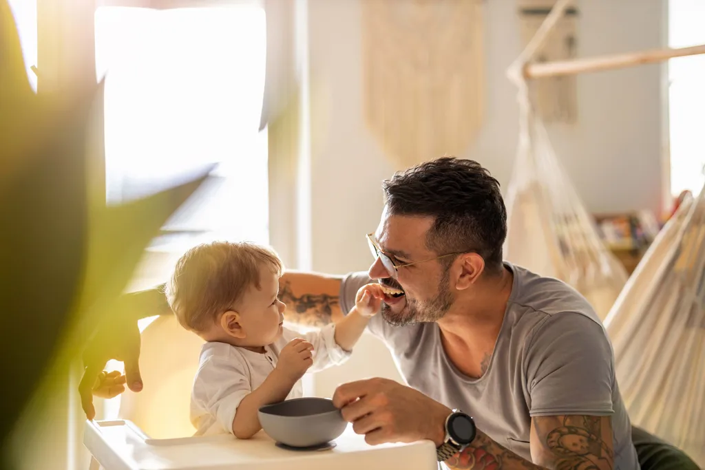 Ein lächelnder Mann mit Tattoos und Brille füttert ein Baby, das in einem Hochstuhl sitzt, in einem sonnendurchfluteten Raum, im Hintergrund ist eine Hängematte zu sehen.