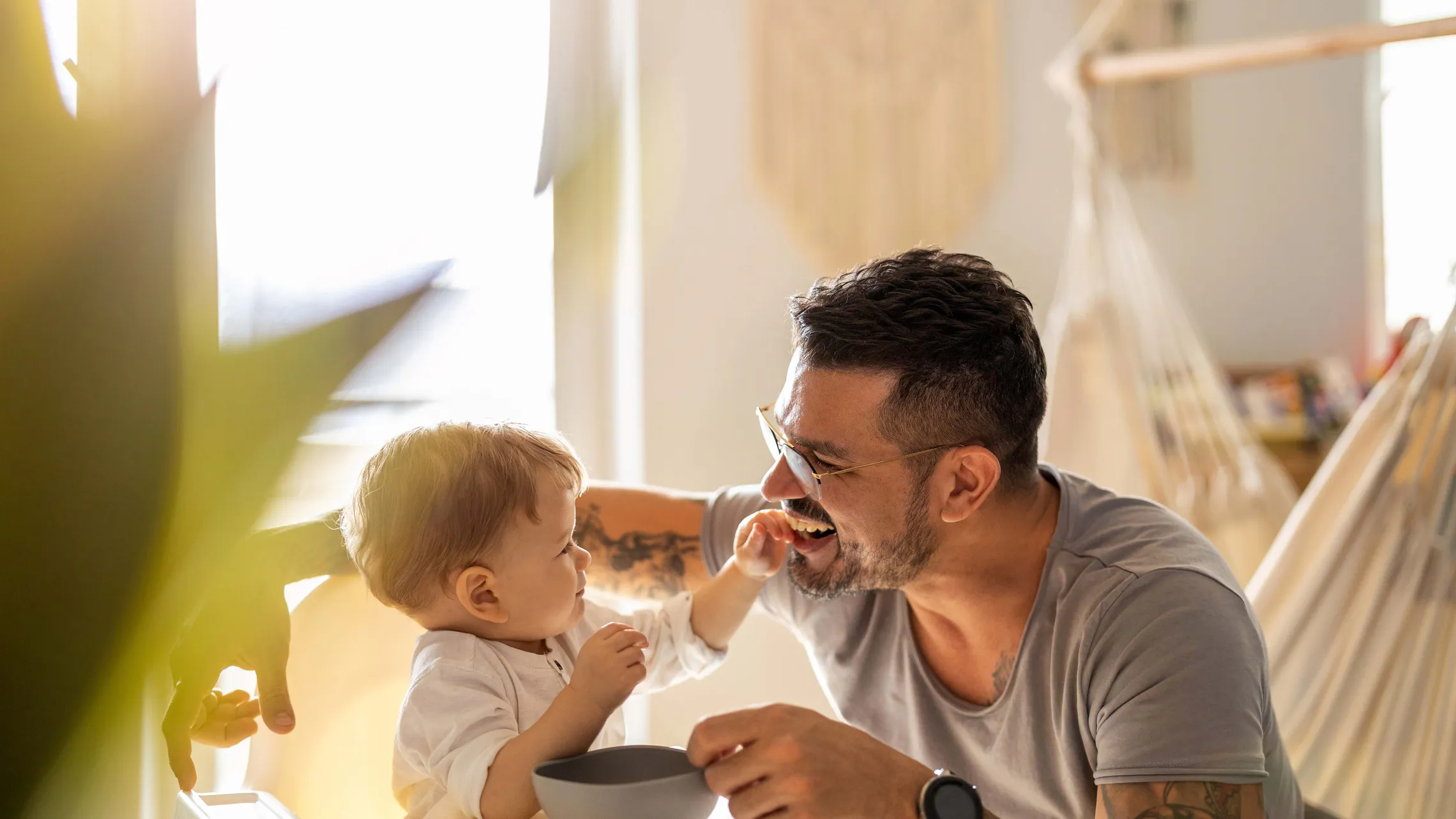 Ein lächelnder Mann mit Tattoos und Brille füttert ein Baby, das in einem Hochstuhl sitzt, in einem sonnendurchfluteten Raum, im Hintergrund ist eine Hängematte zu sehen.