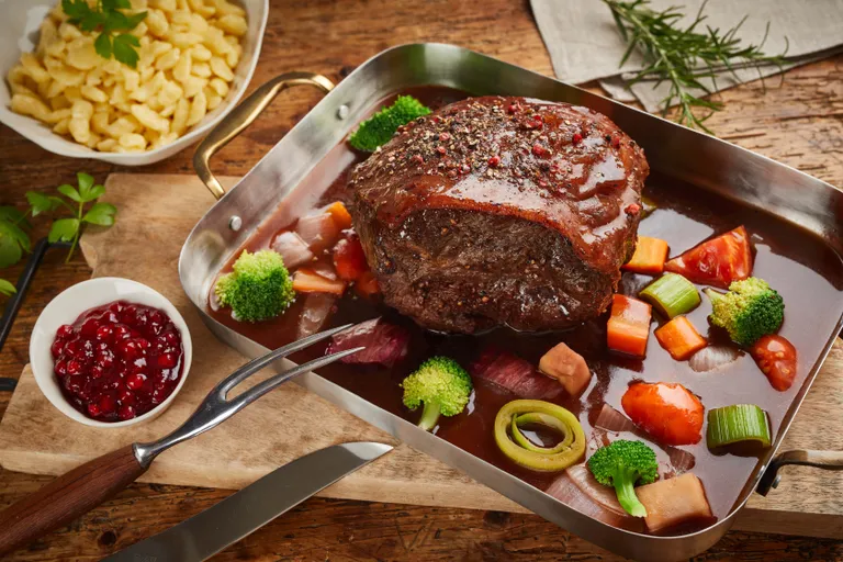 Ein saftiger Rinderbraten in einer Pfanne mit Gemüse und Bratensoße, dazu eine Schüssel mit roter Soße und eine Portion Nudeln, serviert auf einem Holztisch.