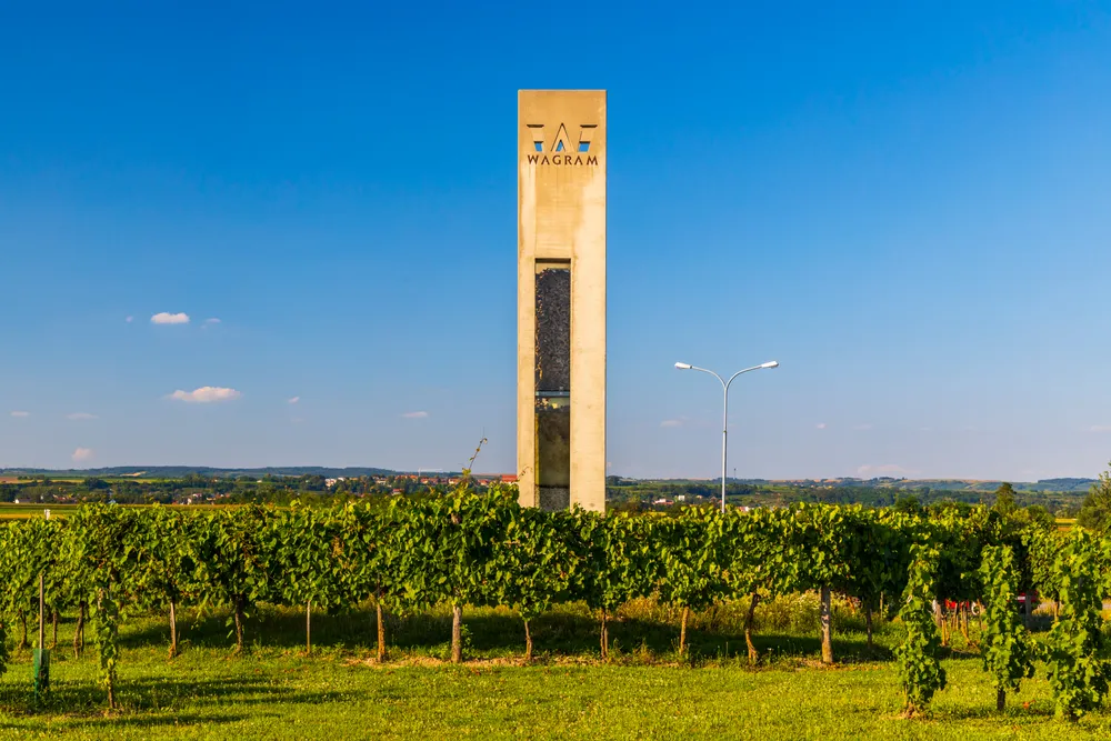 Ein hoher, rechteckiger Steinturm mit der Aufschrift „Wagram“ steht inmitten eines saftig grünen Weinbergs unter einem strahlend blauen Himmel.