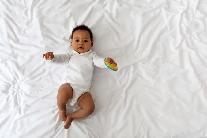 Ein Baby in einem weißen Strampler liegt mit einem entspannten Gesichtsausdruck auf einem Bett und hält ein buntes Spielzeug in der Hand.