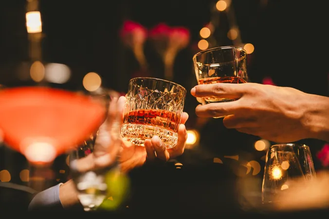 Zwei Hände, die vor einem warmen, verschwommenen Hintergrund mit Bokeh-Lichtern Whiskygläser anstoßen und so eine festliche Atmosphäre schaffen.