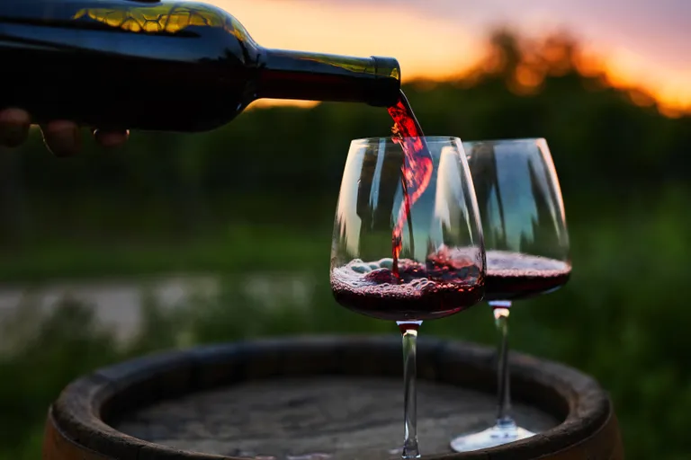 Rotwein wird vor dem Hintergrund eines Sonnenuntergangs aus einem Holzfass in zwei Gläser eingeschenkt.