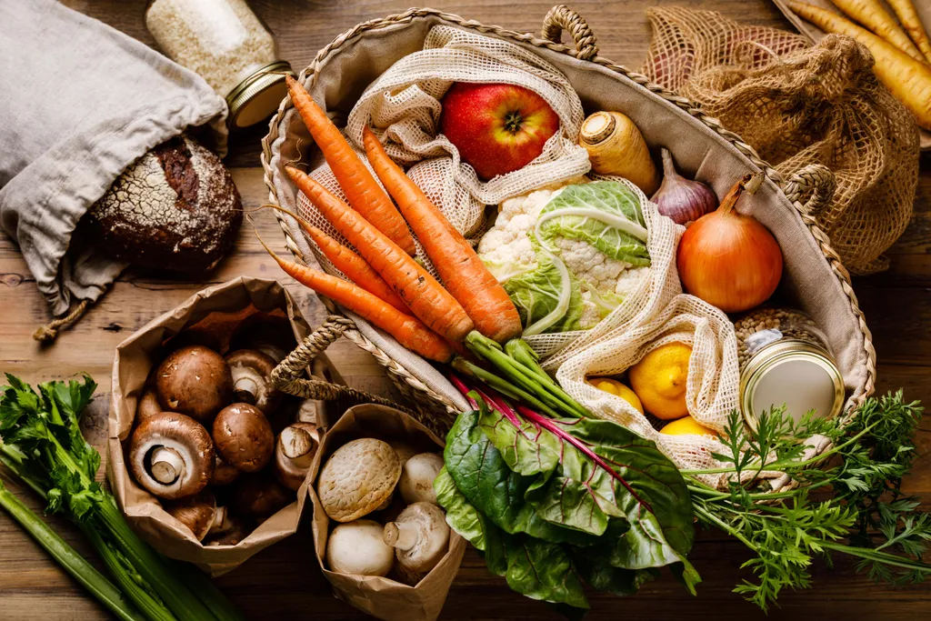 Ein Korb mit frischem Gemüse, darunter Karotten, Blumenkohl und Zwiebeln, umgeben von Pilzen, Sellerie, Brot und einem Glas mit Getreide auf einem Holztisch.