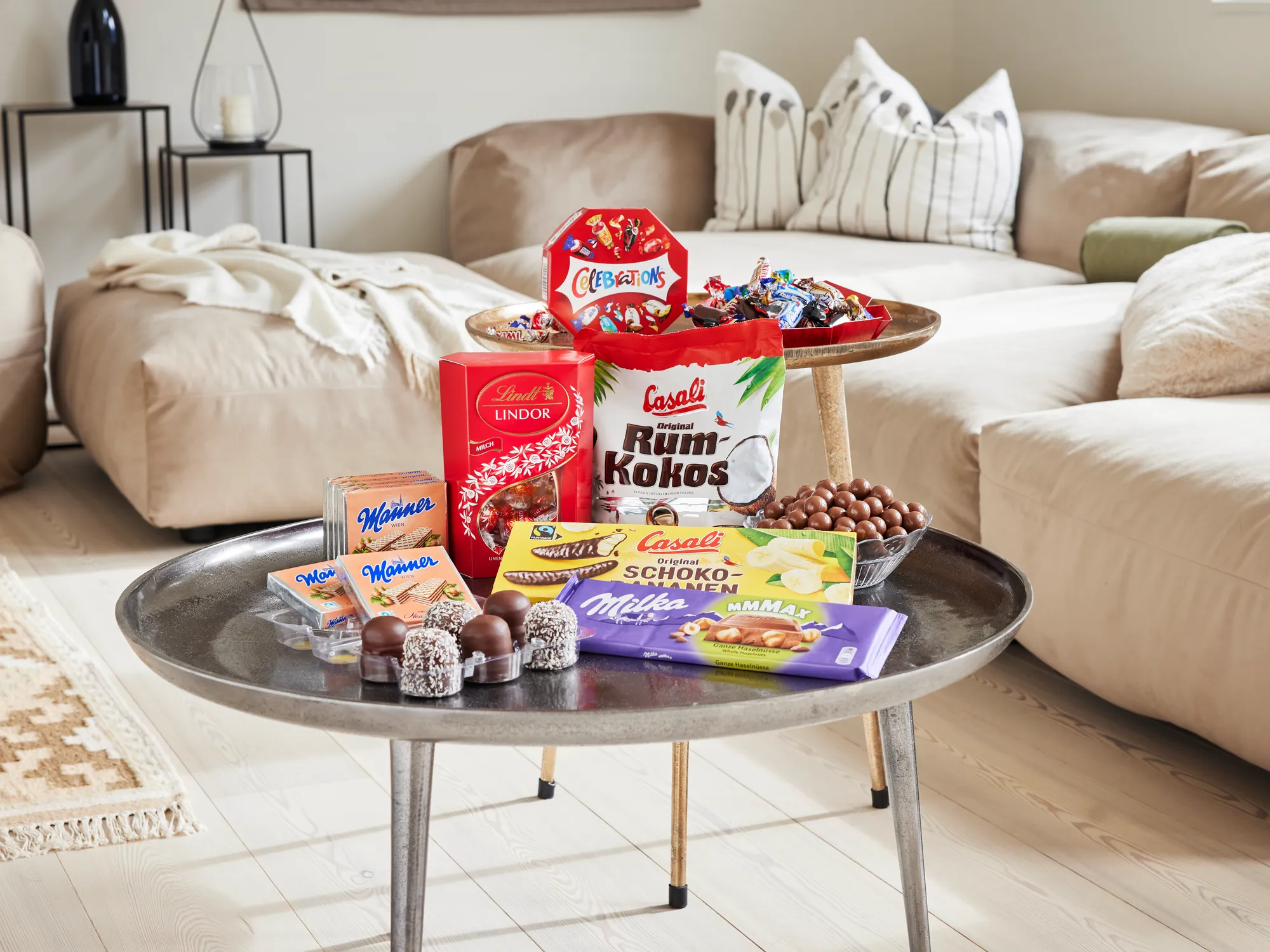 Mehrere verschiedene Süßwaren wie Lindor-Kugeln, Casali Rum-Kokos-Kugeln, Milka Schokolade, Manner-Schnitten und mehr auf einem Couchtisch