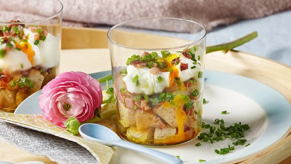 Zwei Gläser mit Toastwürfeln, Ei und Speckwürfeln auf einem Tablett mit Blumen
