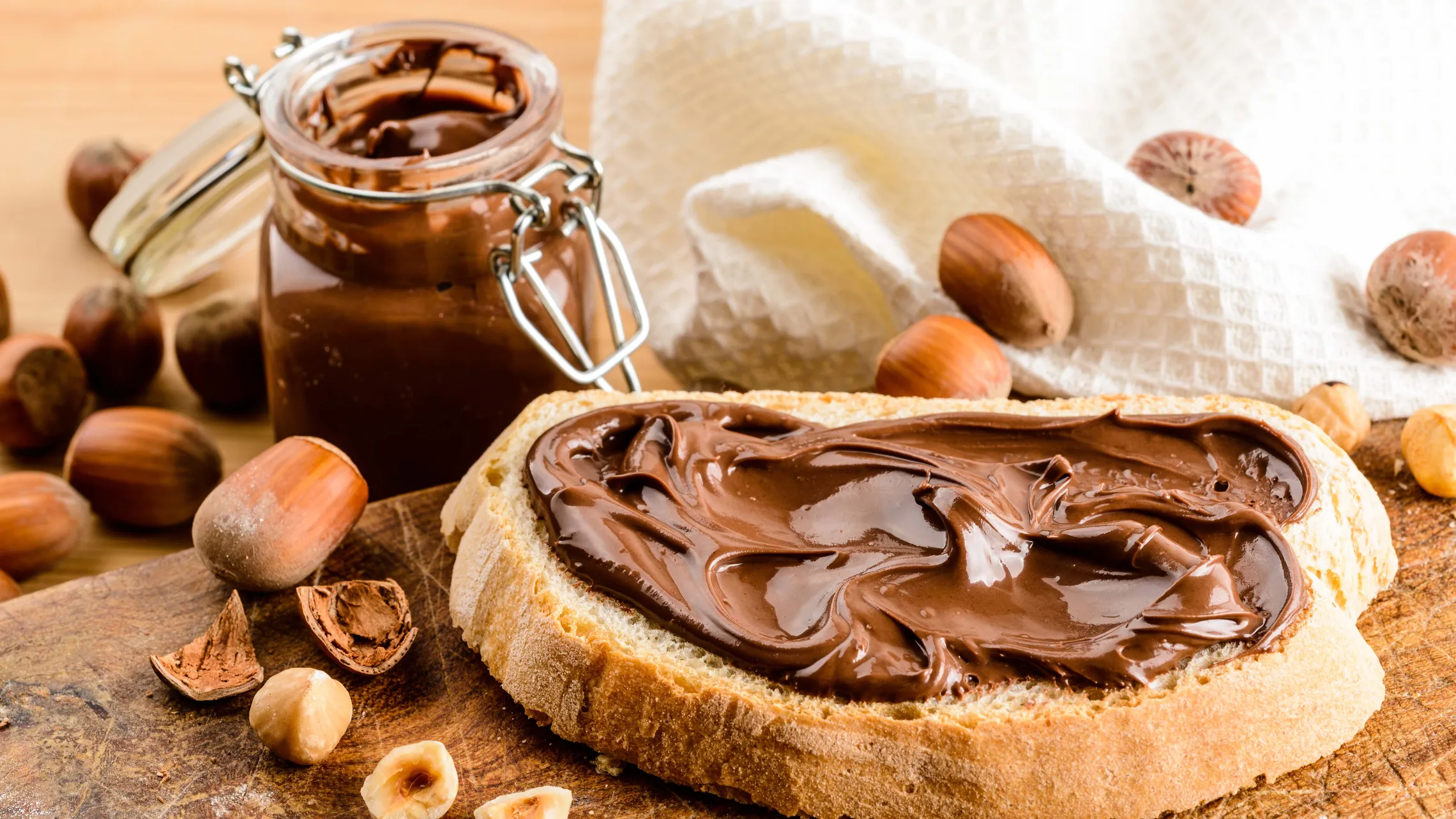 Brotscheibe mit Schokocreme neben einem Glas Schoko-Nuss-Aufstrich und Haselnüssen