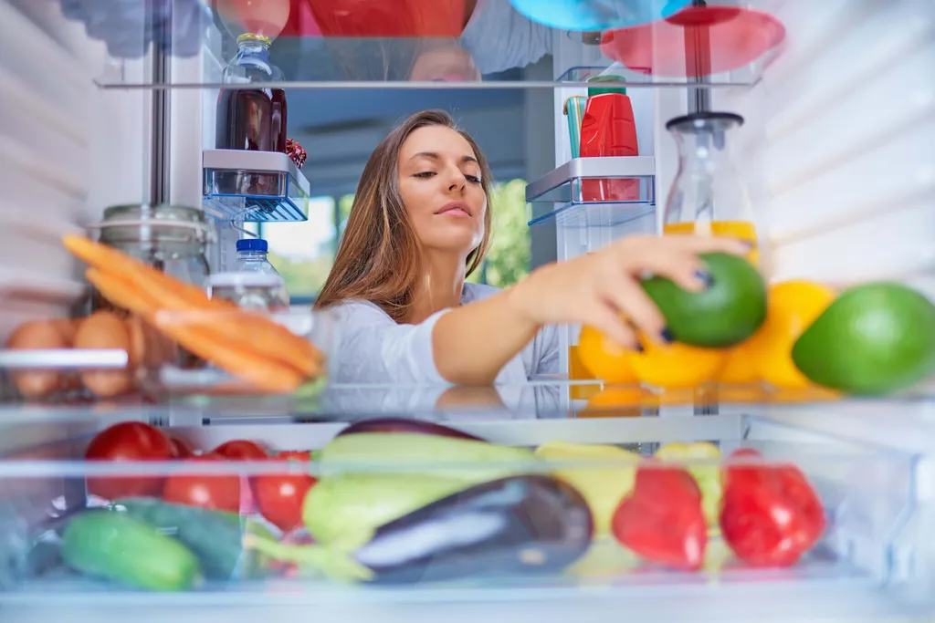 Blick aus dem Kühlschrank auf eine Frau, die frisches Obst und Gemüse einsortiert