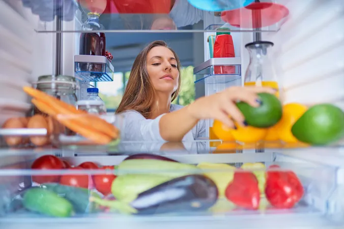 Blick aus dem Kühlschrank auf eine Frau, die frisches Obst und Gemüse einsortiert