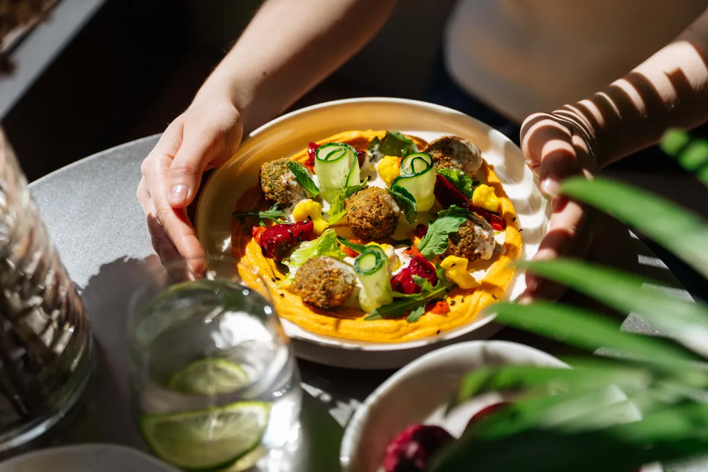 Eine Person hält auf einem Tisch einen bunten Teller mit Falafel, Hummus und Gemüse in der Hand, im Vordergrund steht ein Glas Limettenwasser.