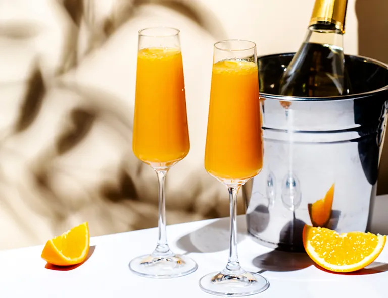 Zwei Mimosa-Cocktails mit Orangensaft und Schaumwein neben Orangenspalten und einer gekühlten Flasche
