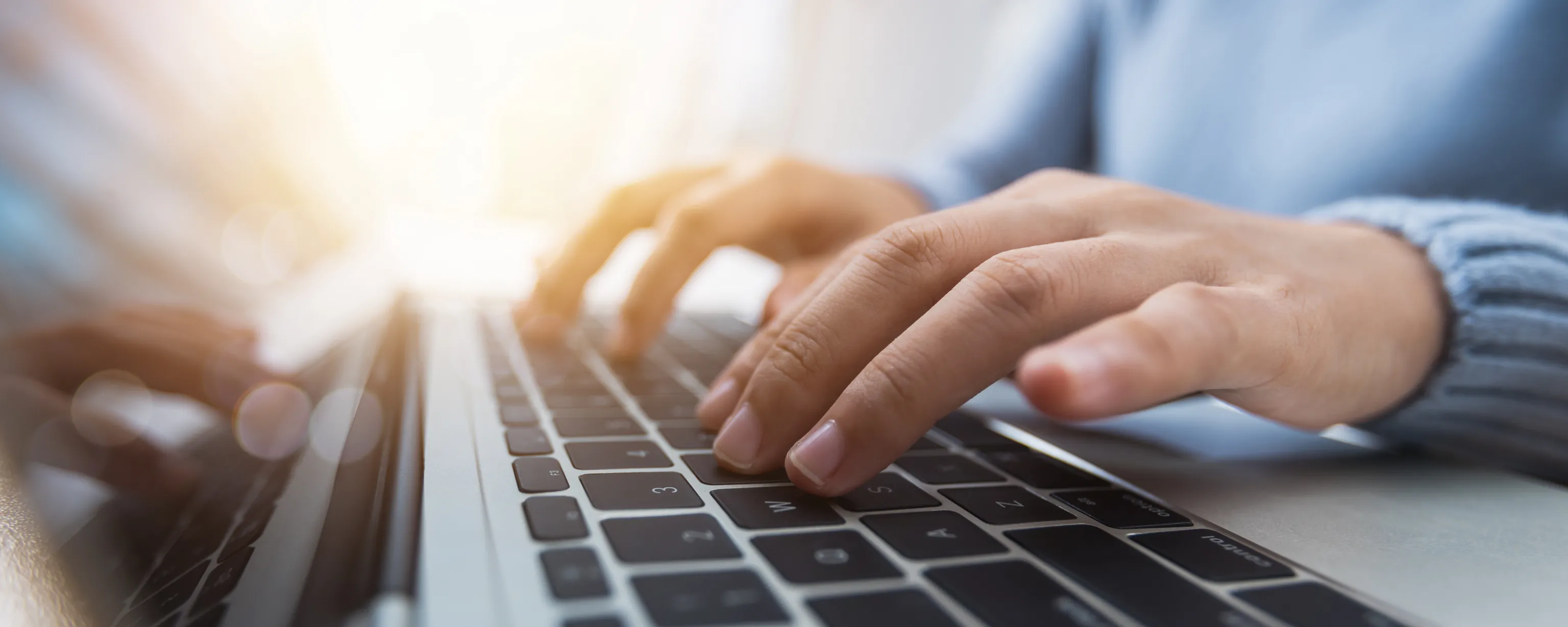 Person schreibt etwas auf dem Laptop, Nahaufnahme des Laptops inklusive Hände und Teil des Oberkörpers, Sonnenstrahlen von hinten