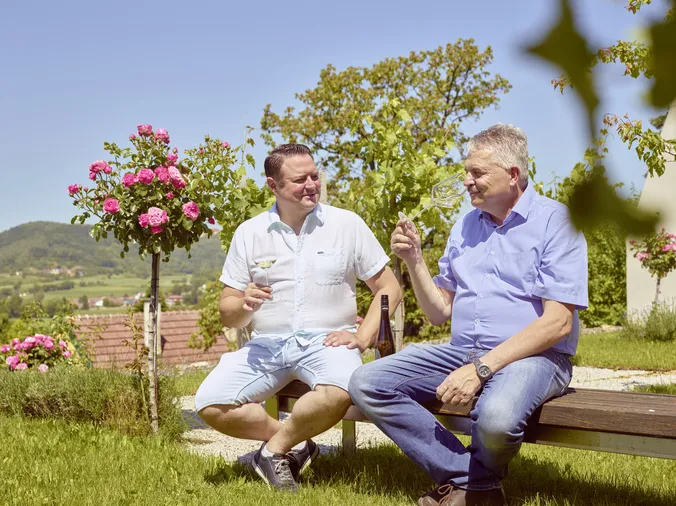 Zwei Männer sitzen draußen auf einer Bank in einem Garten, halten Weingläser in der Hand und sind unter einem strahlend blauen Himmel von blühenden rosa Rosen und Grün umgeben.