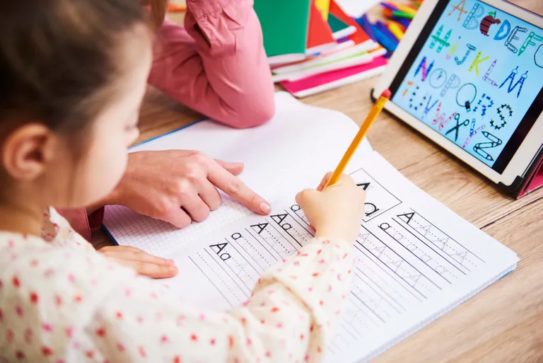 Kind übt das Schreiben von Buchstaben in einem Schreibheft, während eine erwachsene Person unterstützt und ein Tablet das Alphabet zeigt