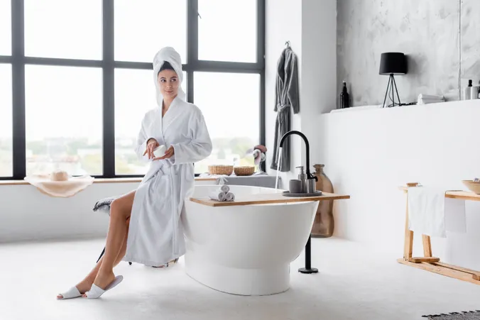 Eine Frau im Bademantel schaut in die Ferne, während sie am Rand ihrer Badewanne sitzt.