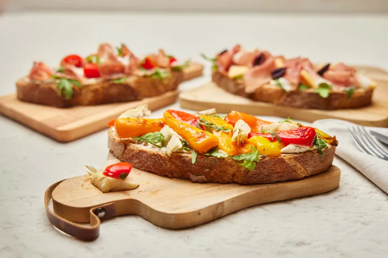 Geröstetes Brot, belegt mit bunten Paprikaschoten, Rucola und Artischocken, auf einem Holzbrett, im Hintergrund sind ähnliche Gerichte verschwommen zu sehen.