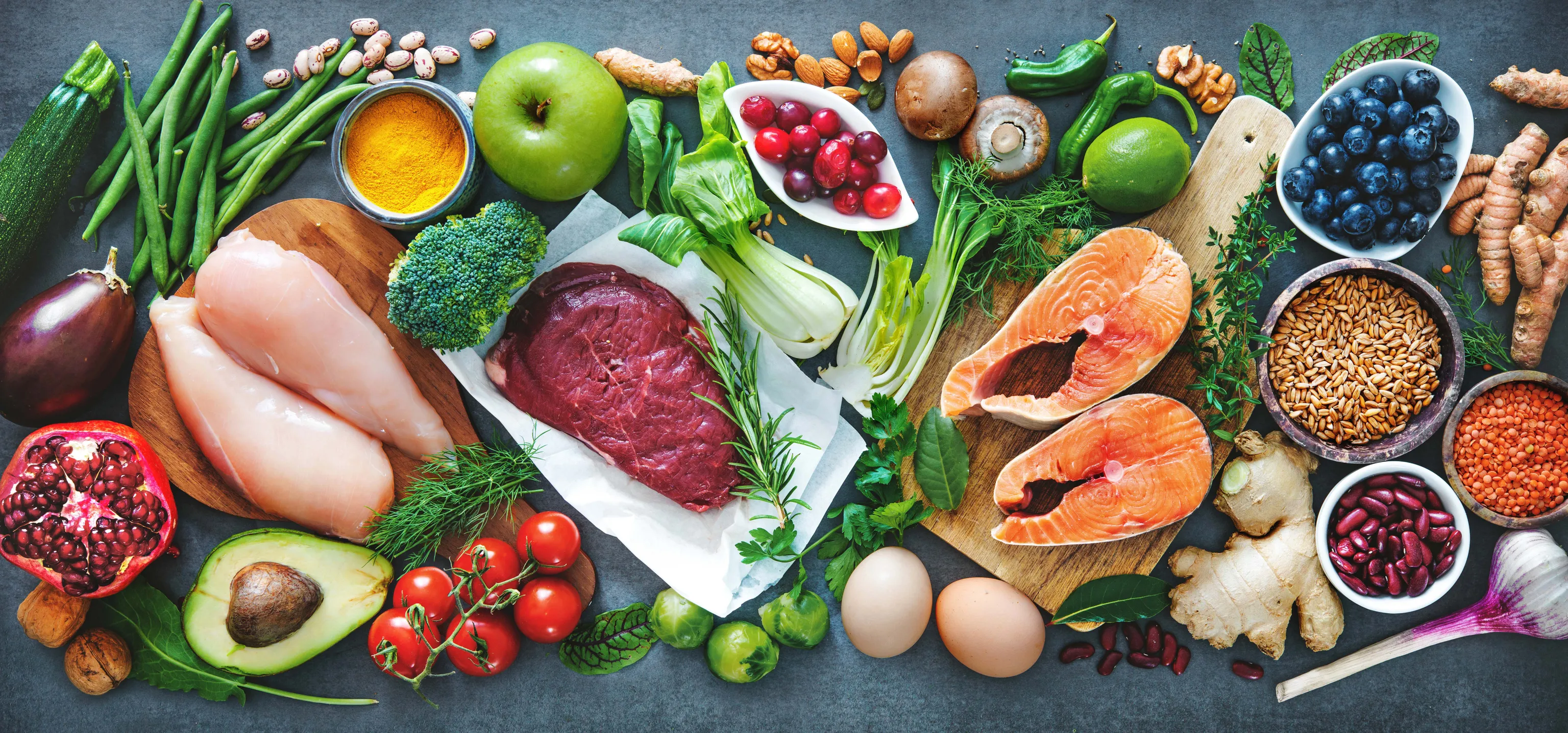 Eine Auswahl an frischen Lebensmitteln, darunter Lachs, Hähnchen, Rindfleisch, Gemüse, Obst, Getreide und Kräuter, die auf einem Tisch angerichtet sind.