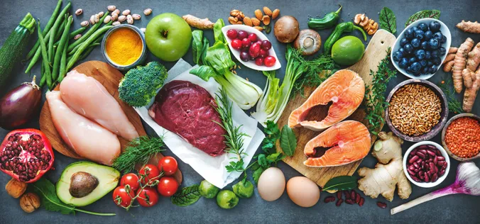 Eine Auswahl an frischen Lebensmitteln, darunter Lachs, Hähnchen, Rindfleisch, Gemüse, Obst, Getreide und Kräuter, die auf einem Tisch angerichtet sind.