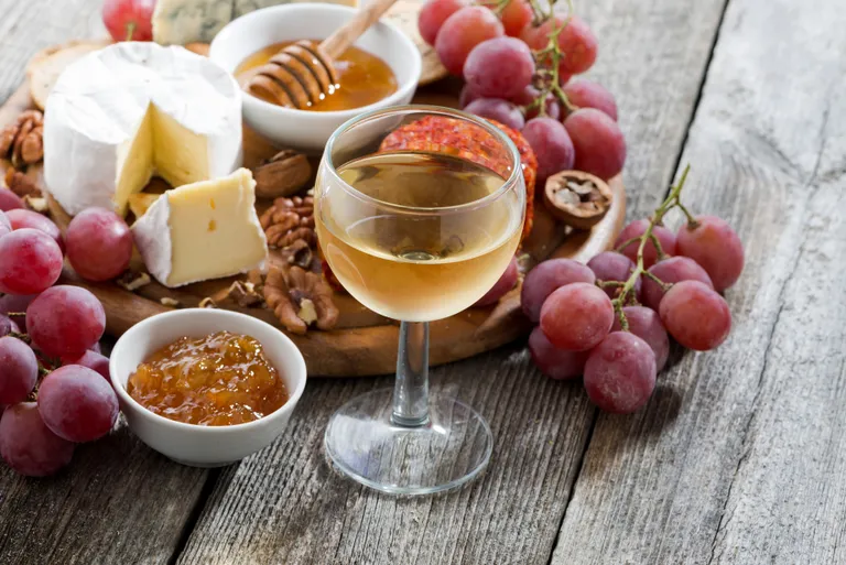 Ein Glas Eiswein, umgeben von Käse, roten Trauben, Honig, Nüssen und Marmelade auf einem rustikalen Holztisch.