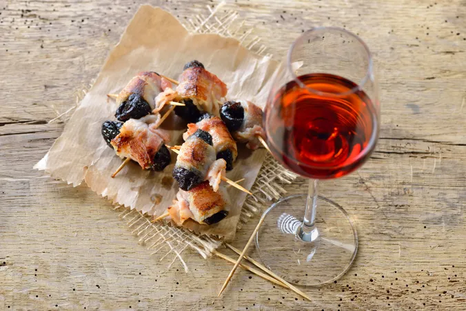 Mit Speck umwickelte Pflaumen am Spieß, auf Backpapier neben einem Glas Rotwein auf einem rustikalen Holztisch.