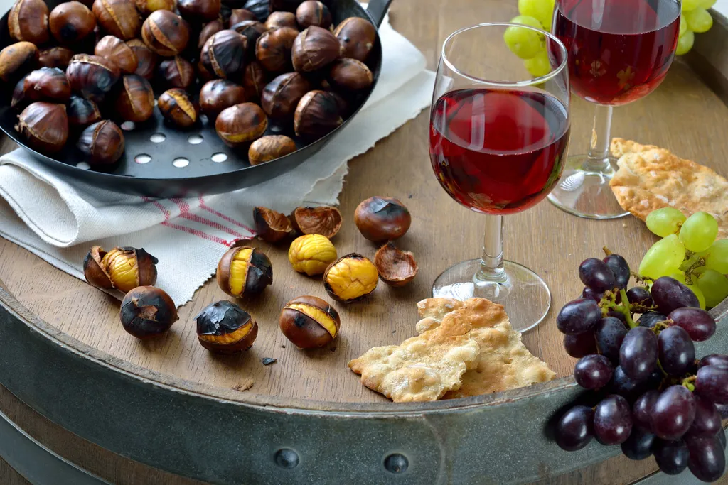 Eine Pfanne mit geröstetetn Maroni und ein Glas Rotweín und Schüttelbrot auf einem Weinfass