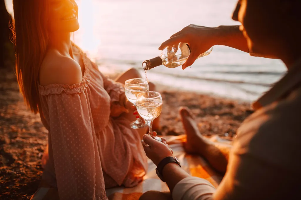Ein Paar am Strand bei Sonnenuntergang; eine Person schenkt Weißwein in zwei Gläser ein und schafft so eine romantische Atmosphäre.