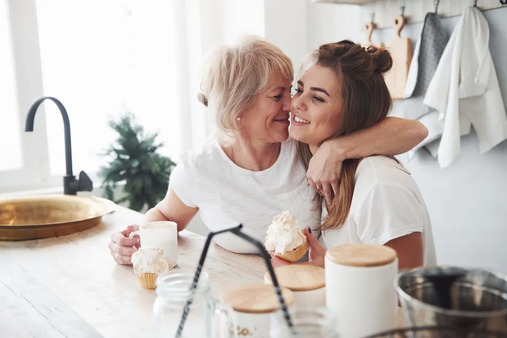 Mutter und erwachsene Tochter umarmen sich in einer Küche, beide essen Cupcakes
