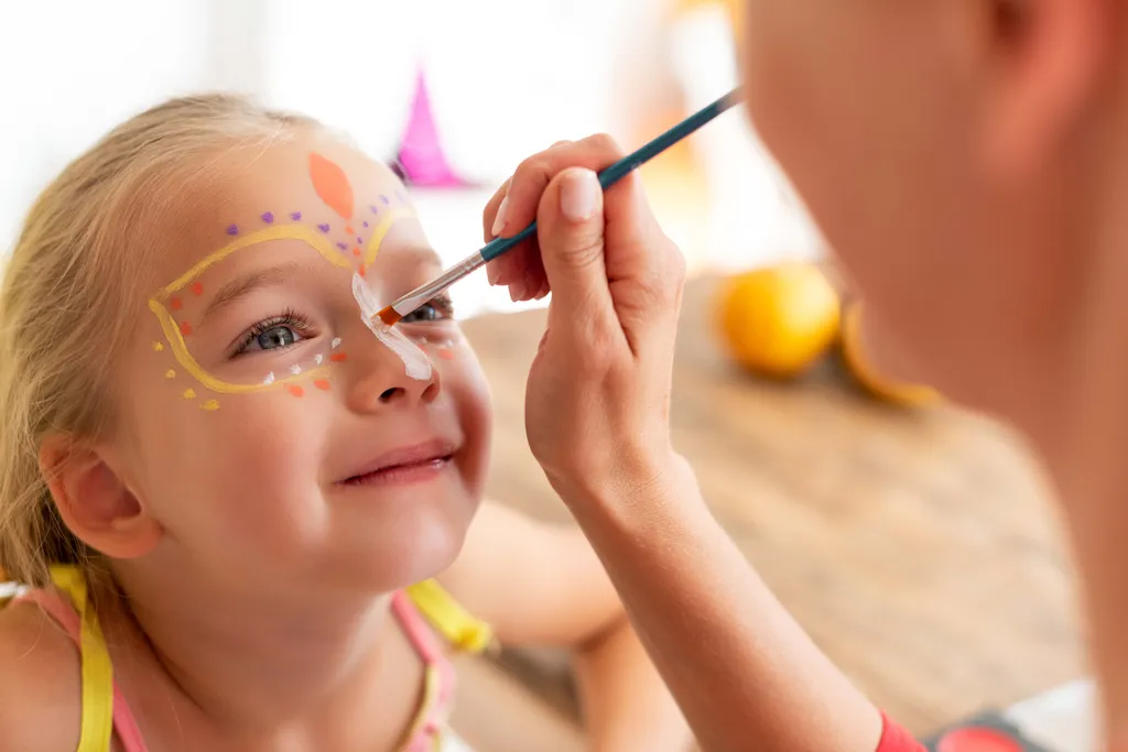 Child with decorative face paint being applied, smiling with eyes closed, indoors on a wooden table.