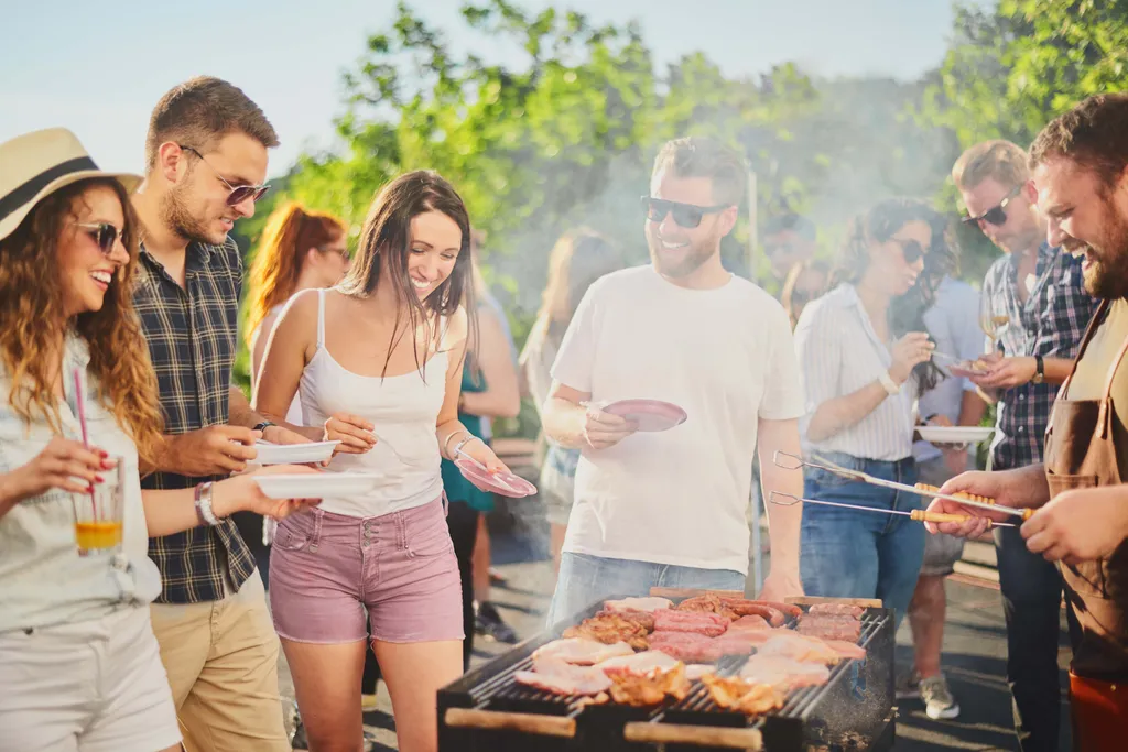 Eine Gruppe von Freunden genießt ein sonniges Grillfest im Freien, hält Teller und Getränke in den Händen, im Vordergrund sind Fleischgrills zu sehen.