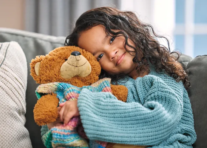 Kind mit blauem Strickpullover mit Teddybär in der Hand, auf der Couch