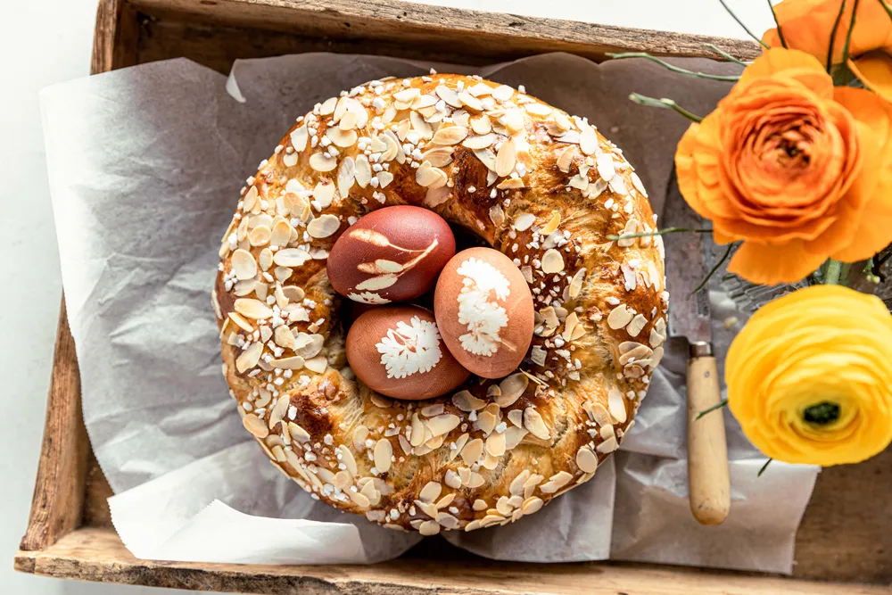 Ein rundes Brot mit Mandelscheiben und drei verzierten Eiern auf einem Holztablett; daneben orangefarbene und gelbe Blumen.