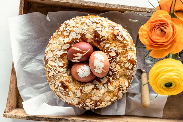 Ein rundes Brot mit Mandelscheiben und drei verzierten Eiern auf einem Holztablett; daneben orangefarbene und gelbe Blumen.