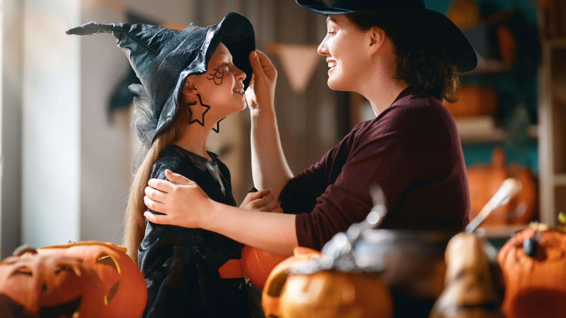 Eine Mutter schminkt ihre Tochter für Halloween, beide tragen einen Hexenhut, im Hintergrunf Kürbislaternen