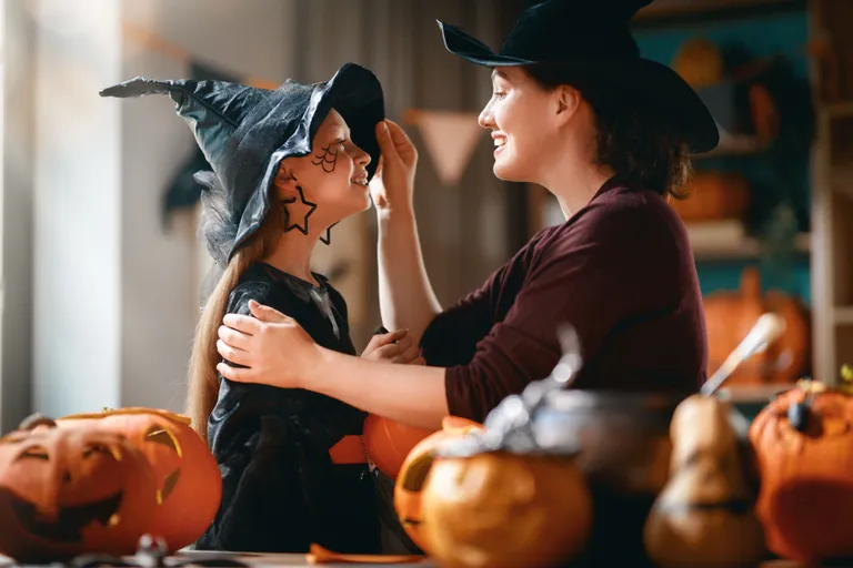 Eine Mutter schminkt ihre Tochter für Halloween, beide tragen einen Hexenhut, im Hintergrunf Kürbislaternen