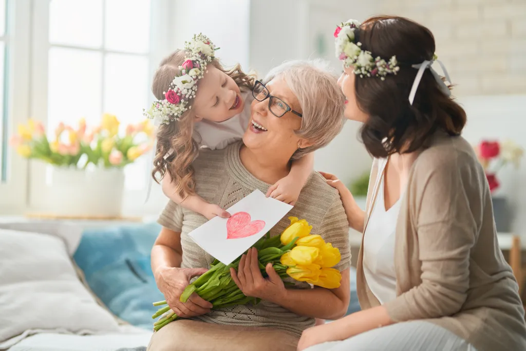 Drei Generationen feiern gemeinsam; eine Großmutter hält Tulpen und eine Karte in der Hand, während sie ihre Tochter und Enkelin umarmt, die Blumenkränze tragen.