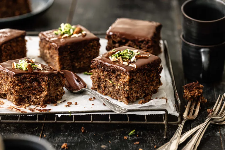 Stücke von Schokoladen-Zucchini-Kuchen mit glänzender Glasur und Nussgarnitur auf einem Tablett, dazu gestapelte schwarze Tassen und Besteck.