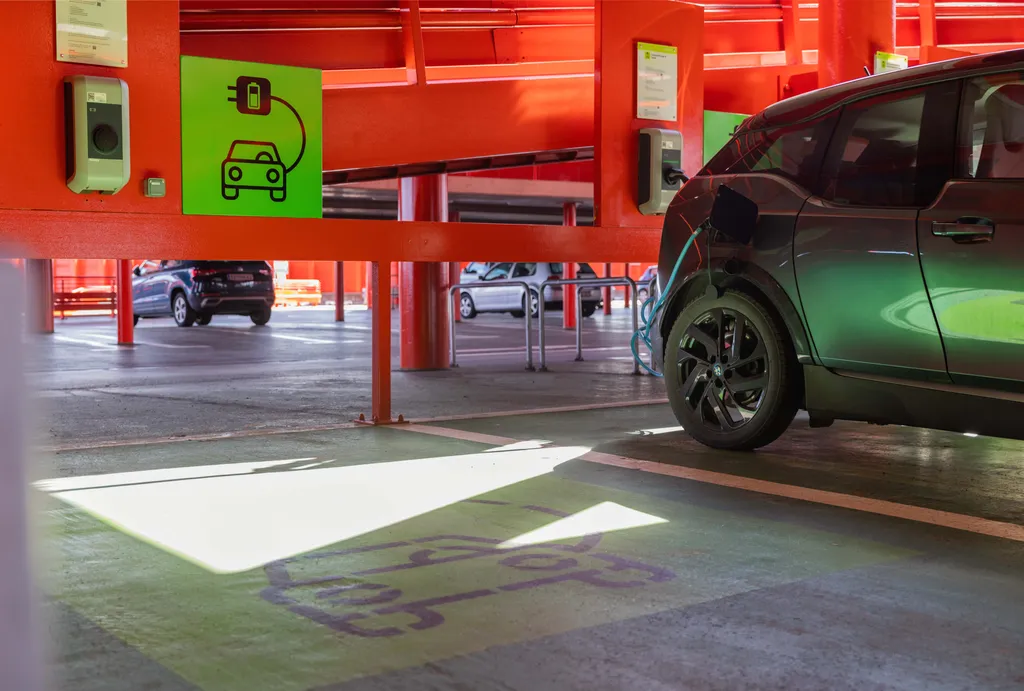 Ein Elektroauto wird in einem leuchtend roten Parkhaus aufgeladen, wobei grüne Schilder auf eine Ladestation hinweisen.