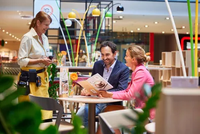 Ein lächelnder Kellner in einer gelben Schürze bedient ein fröhliches Paar in einem modernen Café, das mit lebhaften Dekorationen und Topfpflanzen geschmückt ist.