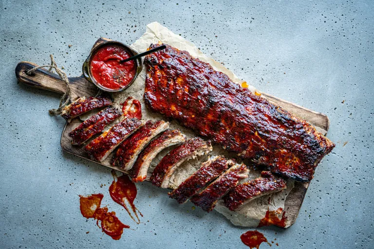 Gegrillte Rippchen auf einem Holzbrett, dazu eine kleine Schale mit roter Sauce, umgeben von Soßenspritzern auf einer strukturierten grauen Oberfläche.