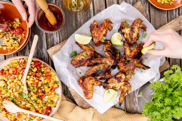 Knusprig gegrillte Hähnchenflügel mit Limettenspalten und buntem Maissalat auf Holztisch