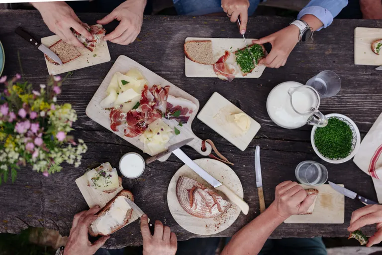 Hände, die auf einem rustikalen Tisch Brot mit Käse, Kräutern und Fleisch zubereiten, umgeben von Butter, Milch und einem Strauß Wildblumen.