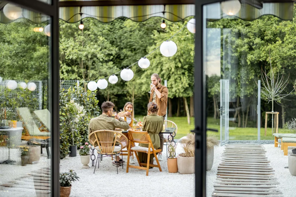 Vier Menschen, die an einem wunderschön gedeckten Tisch im Freien eine Mahlzeit genießen, umgeben von Grün und Laternen – aufgenommen durch große Glastüren.