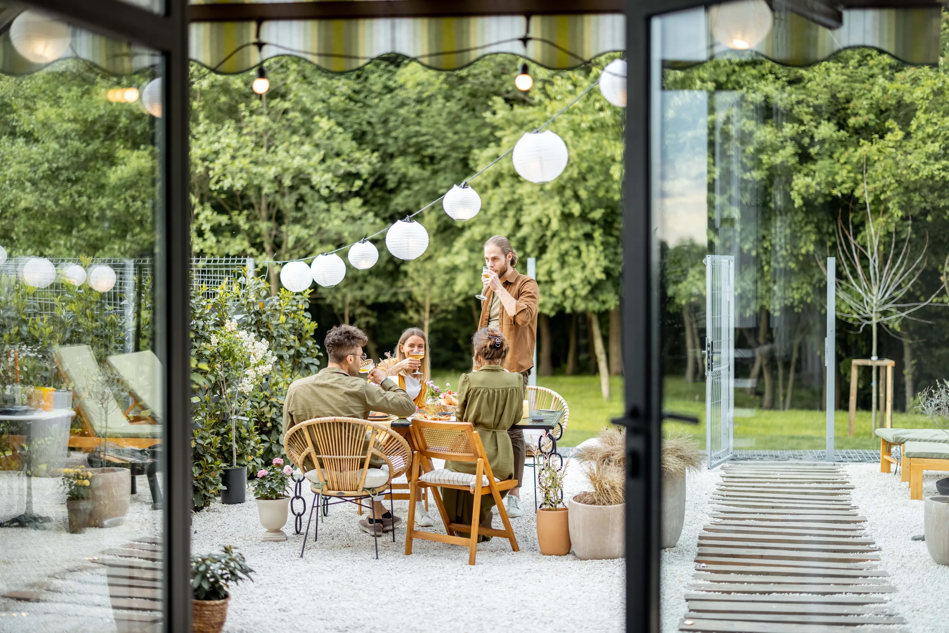 Vier Menschen, die an einem wunderschön gedeckten Tisch im Freien eine Mahlzeit genießen, umgeben von Grün und Laternen – aufgenommen durch große Glastüren.