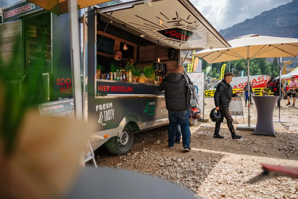 Szene am Foodtruck, in der ein Mann bestellt, während ein anderer in Motorradkleidung daneben steht. Außenbereich mit Sonnenschirmen und Bergkulisse.
