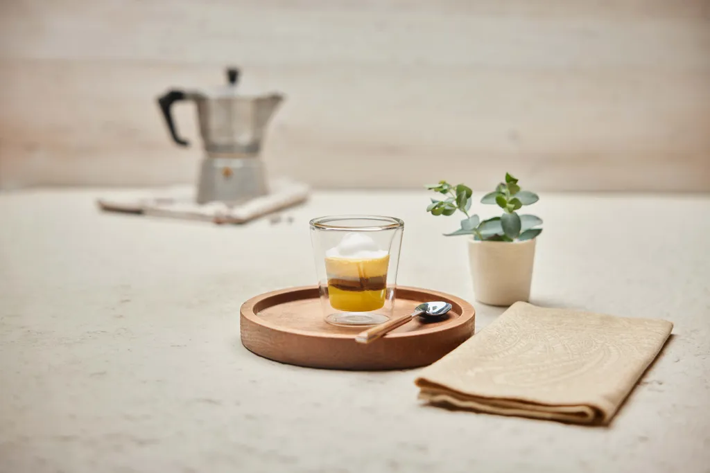 Ein Glas mit geschichtetem Espresso und Milchschaum auf einem Holztablett, im Hintergrund ein Löffel, eine Serviette, eine Pflanze und eine Mokkakanne.