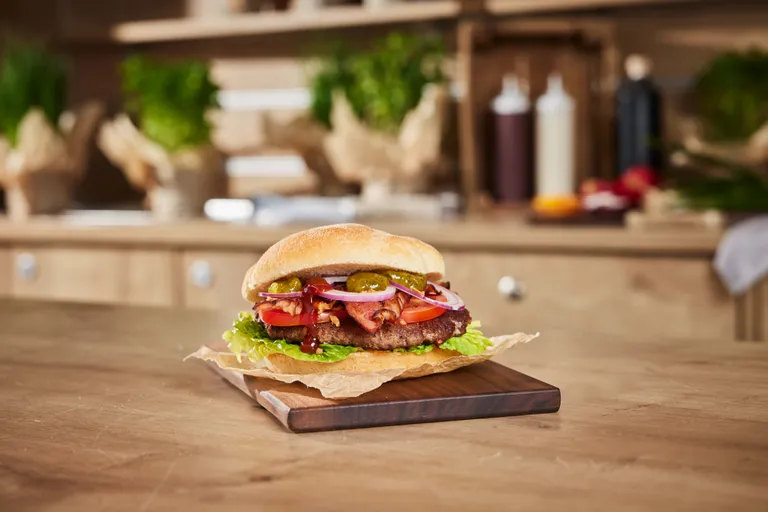 Ein reichhaltig belegter Burger mit Salat, Speck, Zwiebeln, Gurken und Tomate auf einem Holzbrett in einer rustikalen Küchenumgebung.