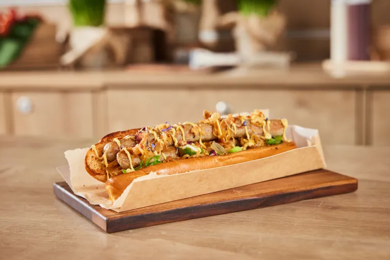 Ein Gourmet-Hotdog mit Zwiebeln, Senf und Kräutern, serviert auf einem Holzbrett mit Backpapier.