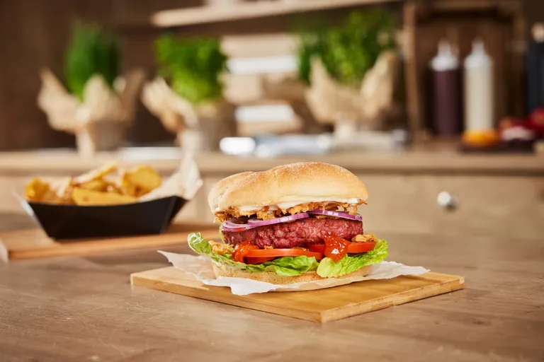 Ein saftiger Burger mit Salat, Tomate und Zwiebeln auf einem Holzbrett, im Hintergrund dazu dick geschnittene Pommes frites.